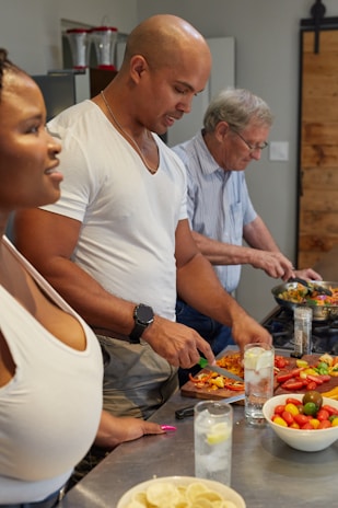 Three people are engaged in preparing food in a kitchen. They are cutting vegetables on a countertop scattered with colorful peppers and tomatoes. Two glasses of a clear beverage with lemon slices are nearby, along with a bowl of cherry tomatoes.