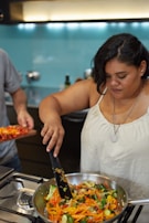 A woman is cooking in a modern kitchen, stirring a pan filled with a colorful mixture of sliced vegetables. The stovetop and kitchen appliances are visible, with a light blue backsplash and warm lighting.