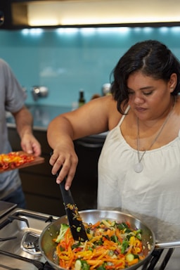 A woman is cooking in a modern kitchen, stirring a pan filled with a colorful mixture of sliced vegetables. The stovetop and kitchen appliances are visible, with a light blue backsplash and warm lighting.