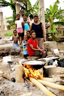 A group of children and two adults are gathered outdoors, smiling at the camera. They are surrounded by greenery with banana trees in the background. In the foreground, there's an open fire with pots placed on it, suggesting a cooking activity. The setting appears to be informal and communal.