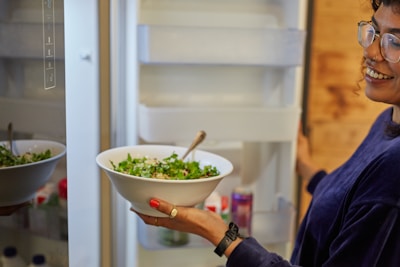 A person holding a bowl filled with a fresh green salad is standing in front of an open refrigerator. The person is smiling and has curly hair and glasses, wearing a dark sweater. The refrigerator door is slightly open, revealing several containers and bottles inside.