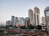 A modern cityscape featuring a series of tall skyscrapers with diverse architectural designs. Each building exhibits a unique style, ranging from glass-covered structures to more traditional concrete designs. A railway with tracks and a platform is visible in the foreground, with people waiting for trains. The scene captures both urban development and daily transit activity.