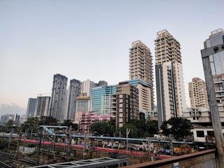 A modern cityscape featuring a series of tall skyscrapers with diverse architectural designs. Each building exhibits a unique style, ranging from glass-covered structures to more traditional concrete designs. A railway with tracks and a platform is visible in the foreground, with people waiting for trains. The scene captures both urban development and daily transit activity.