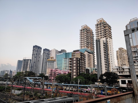 A modern cityscape featuring a series of tall skyscrapers with diverse architectural designs. Each building exhibits a unique style, ranging from glass-covered structures to more traditional concrete designs. A railway with tracks and a platform is visible in the foreground, with people waiting for trains. The scene captures both urban development and daily transit activity.