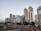 A modern cityscape featuring a series of tall skyscrapers with diverse architectural designs. Each building exhibits a unique style, ranging from glass-covered structures to more traditional concrete designs. A railway with tracks and a platform is visible in the foreground, with people waiting for trains. The scene captures both urban development and daily transit activity.