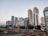 A modern cityscape featuring a series of tall skyscrapers with diverse architectural designs. Each building exhibits a unique style, ranging from glass-covered structures to more traditional concrete designs. A railway with tracks and a platform is visible in the foreground, with people waiting for trains. The scene captures both urban development and daily transit activity.