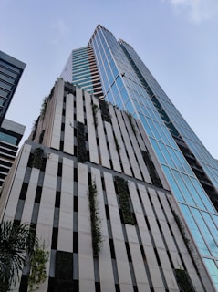 A modern high-rise building with sleek glass panels reflecting the sky. Vertical sections of the facade are interspersed with greenery, giving it a natural touch. The building's design features a mix of vertical and horizontal lines, creating a dynamic urban aesthetic.