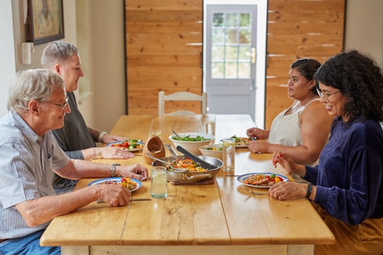 A group of four people sits around a wooden dining table, sharing a meal. Different plates of colorful food, as well as a frying pan with assorted vegetables, are arranged in front of them. The setting is bright and homey, with wooden paneling and a door in the background. Everyone appears engaged in conversation and enjoying the gathering.