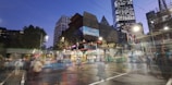 Urban scene featuring modern architecture with a blend of illuminated skyscrapers and busy streets. A tram or bus is in motion, and pedestrians are visible even as they appear slightly blurred. The sky is transitioning to evening hues, and artificial lights brighten the scene.