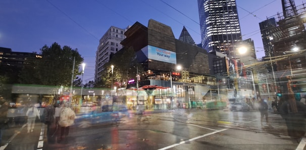 Urban scene featuring modern architecture with a blend of illuminated skyscrapers and busy streets. A tram or bus is in motion, and pedestrians are visible even as they appear slightly blurred. The sky is transitioning to evening hues, and artificial lights brighten the scene.