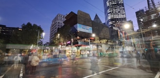 Urban scene featuring modern architecture with a blend of illuminated skyscrapers and busy streets. A tram or bus is in motion, and pedestrians are visible even as they appear slightly blurred. The sky is transitioning to evening hues, and artificial lights brighten the scene.