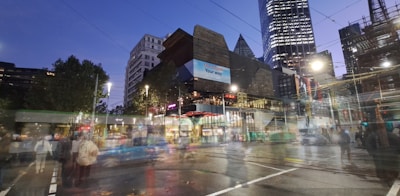 Urban scene featuring modern architecture with a blend of illuminated skyscrapers and busy streets. A tram or bus is in motion, and pedestrians are visible even as they appear slightly blurred. The sky is transitioning to evening hues, and artificial lights brighten the scene.