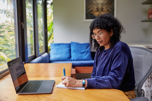 A person wearing glasses and a blue long-sleeve shirt is sitting at a wooden table, writing in a notebook with a pen. They are seated on a gray office chair beside a laptop showing a webpage. Behind them, there are blue cushions on a couch, a large window with greenery outside, and a wall with a framed picture.