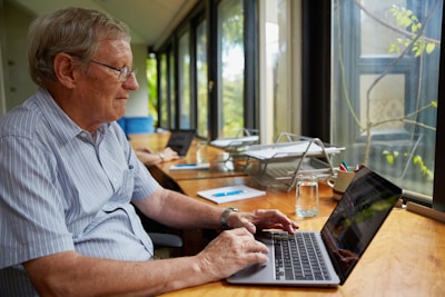 An older man is using a laptop at a wooden desk near a large window. The room has natural lighting and there are glass cups, notebooks, and a pen holder with colored pens on the desk. Another person in the background is also working on a laptop.
