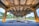 An architectural structure with large concrete beams and pillars in a landscaped outdoor setting. The area is illuminated with warm lights, highlighting the design elements. Trees and greenery are visible in the background, contrasting with the industrial feel of the framework.