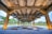 An architectural structure with large concrete beams and pillars in a landscaped outdoor setting. The area is illuminated with warm lights, highlighting the design elements. Trees and greenery are visible in the background, contrasting with the industrial feel of the framework.