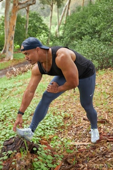 A man stands outdoors on a grassy patch, stretching by placing one foot on a tree stump. He is wearing a black tank top, blue leggings, white sneakers, and a backward blue cap. The background features greenery and tall trees.