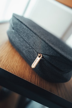 A close-up view of a dark gray fabric pouch placed on a wooden surface. The pouch has a gold zipper pull with the brand name 'SONY' engraved on it. The background is softly blurred, highlighting the texture and detail of the pouch and table.