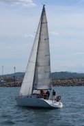 A sailboat with a large mast glides on the water, manned by three individuals. The backdrop features a rocky shoreline and distant hills under a partly cloudy sky.