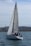 A sailboat with a large mast glides on the water, manned by three individuals. The backdrop features a rocky shoreline and distant hills under a partly cloudy sky.