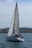 A sailboat with a large mast glides on the water, manned by three individuals. The backdrop features a rocky shoreline and distant hills under a partly cloudy sky.