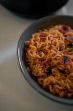 A bowl filled with cooked instant noodles, garnished with narutomaki, which are small, round slices with a decorative swirl or character printed on them. The noodles have a slightly glossy texture, indicating they may be coated with sauce. A few pieces of green vegetable like spinach or seaweed are visible among the noodles.