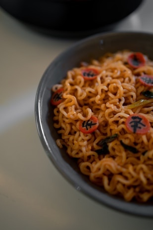 A bowl filled with cooked instant noodles, garnished with narutomaki, which are small, round slices with a decorative swirl or character printed on them. The noodles have a slightly glossy texture, indicating they may be coated with sauce. A few pieces of green vegetable like spinach or seaweed are visible among the noodles.
