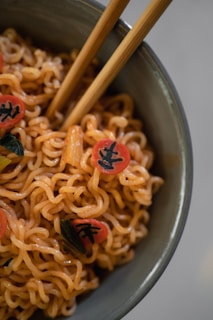 A close-up of a bowl filled with cooked noodles garnished with red circle-shaped toppings featuring black markings. The noodles are glossy and intertwined, with a pair of wooden chopsticks resting within the bowl. The dish appears to have some green ingredients mixed in.