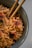 A close-up of a bowl filled with cooked noodles garnished with red circle-shaped toppings featuring black markings. The noodles are glossy and intertwined, with a pair of wooden chopsticks resting within the bowl. The dish appears to have some green ingredients mixed in.