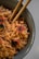 A close-up of a bowl filled with cooked noodles garnished with red circle-shaped toppings featuring black markings. The noodles are glossy and intertwined, with a pair of wooden chopsticks resting within the bowl. The dish appears to have some green ingredients mixed in.