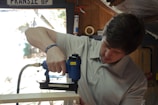 A person using a blue pneumatic nail gun to work on a piece of wood in a workshop setting. The individual is wearing a light gray shirt and a wristband. The workshop has wooden walls and various tools can be seen in the background.