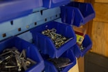 A collection of blue plastic bins mounted on a wall, containing various hardware items such as screws, nails, and hooks. The bins are organized, giving a tidy appearance, with some items packaged in plastic.
