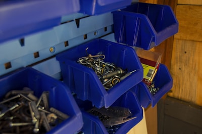A collection of blue plastic bins mounted on a wall, containing various hardware items such as screws, nails, and hooks. The bins are organized, giving a tidy appearance, with some items packaged in plastic.