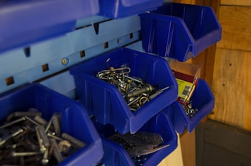 A collection of blue plastic bins mounted on a wall, containing various hardware items such as screws, nails, and hooks. The bins are organized, giving a tidy appearance, with some items packaged in plastic.