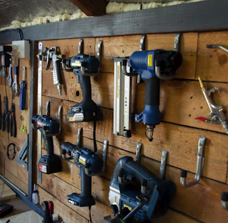 A well-organized workshop scene displays various power tools and hand tools neatly hung on a wooden wall. The tools include drills, screwdrivers, pliers, and wrenches, among others. The background is made of wooden planks, creating a rustic ambiance.
