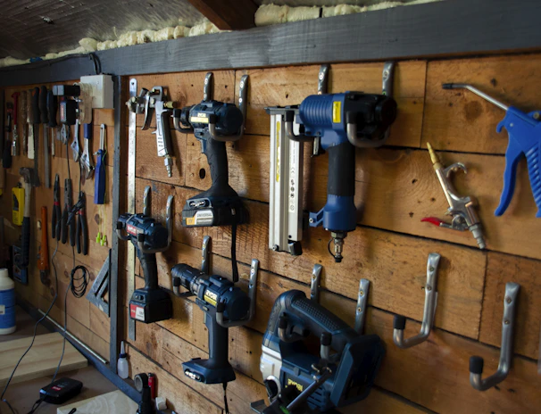 A well-organized workshop scene displays various power tools and hand tools neatly hung on a wooden wall. The tools include drills, screwdrivers, pliers, and wrenches, among others. The background is made of wooden planks, creating a rustic ambiance.