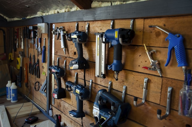 A well-organized workshop scene displays various power tools and hand tools neatly hung on a wooden wall. The tools include drills, screwdrivers, pliers, and wrenches, among others. The background is made of wooden planks, creating a rustic ambiance.