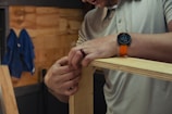 A person wearing a grey polo shirt is assembling wooden boards, focused on their hands holding the panels together. They wear an orange wristwatch and a ring. A blue cloth hangs on the wooden wall in the background.