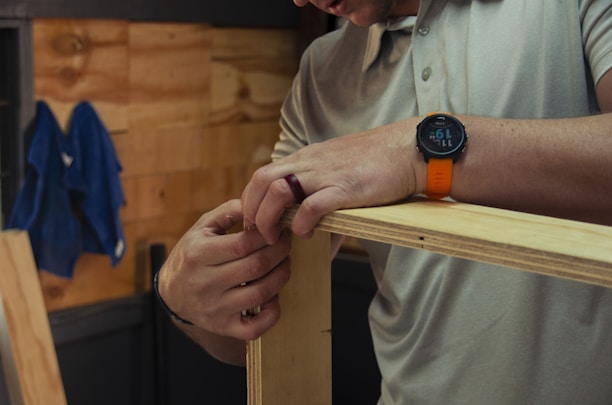 A person wearing a grey polo shirt is assembling wooden boards, focused on their hands holding the panels together. They wear an orange wristwatch and a ring. A blue cloth hangs on the wooden wall in the background.