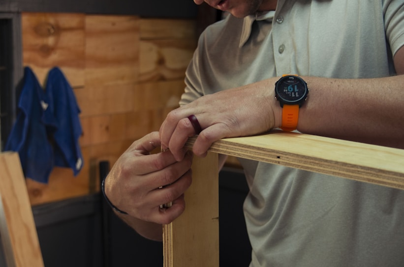 A person wearing a grey polo shirt is assembling wooden boards, focused on their hands holding the panels together. They wear an orange wristwatch and a ring. A blue cloth hangs on the wooden wall in the background.