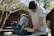A person is operating a Ryobi chop saw outdoors, focusing on cutting a piece of wood. The setting includes trees with bare branches and a brick wall in the background, indicating a backyard or garden workspace. The individual is casually dressed, suggesting a DIY project or home maintenance.