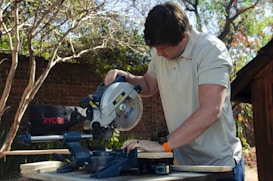 A person is operating a Ryobi chop saw outdoors, focusing on cutting a piece of wood. The setting includes trees with bare branches and a brick wall in the background, indicating a backyard or garden workspace. The individual is casually dressed, suggesting a DIY project or home maintenance.