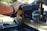 A close-up of a person using a power miter saw to cut a piece of wood. The saw has a blue casing with visible logos and warnings, and it is set on a workbench outdoors. The person’s hand is holding the handle for guidance and control.