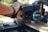 A close-up of a person using a power miter saw to cut a piece of wood. The saw has a blue casing with visible logos and warnings, and it is set on a workbench outdoors. The person’s hand is holding the handle for guidance and control.