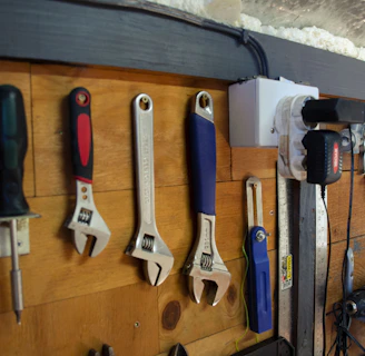 A wooden wall with an assortment of tools neatly hung on hooks. The tools include a screwdriver, several adjustable wrenches, a measuring tape, a utility knife, a drill, and an air compressor. The setup is organized and appears to be part of a workshop environment.