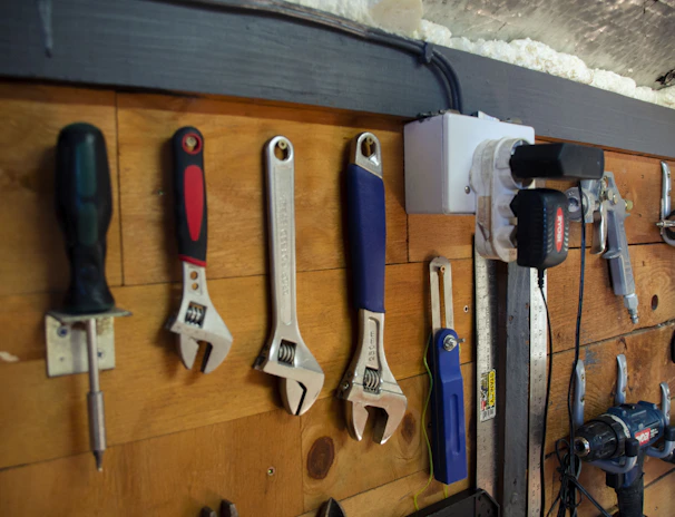 A wooden wall with an assortment of tools neatly hung on hooks. The tools include a screwdriver, several adjustable wrenches, a measuring tape, a utility knife, a drill, and an air compressor. The setup is organized and appears to be part of a workshop environment.