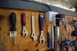 A wooden wall with an assortment of tools neatly hung on hooks. The tools include a screwdriver, several adjustable wrenches, a measuring tape, a utility knife, a drill, and an air compressor. The setup is organized and appears to be part of a workshop environment.