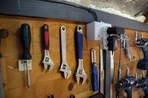 A wooden wall with an assortment of tools neatly hung on hooks. The tools include a screwdriver, several adjustable wrenches, a measuring tape, a utility knife, a drill, and an air compressor. The setup is organized and appears to be part of a workshop environment.