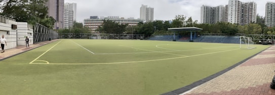 A large artificial turf sports field surrounded by a high fence, located in an urban area with tall residential buildings and trees. A small covered seating area is visible on one side, and there is a soccer goalpost positioned at one end. Two people are walking along the path on the left side of the field.