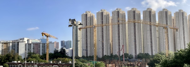 A line of tall residential buildings stands prominently against a clear blue sky. In the foreground, multiple construction cranes are visible, suggesting on-going development. The area is surrounded by lush greenery and other smaller buildings.